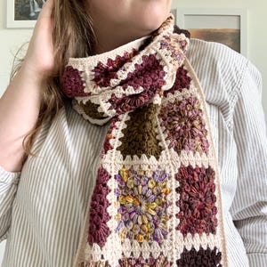 May include: A crocheted scarf with a multi-colored granny square pattern in shades of brown, pink, and green. The scarf is made with a cream-colored yarn.