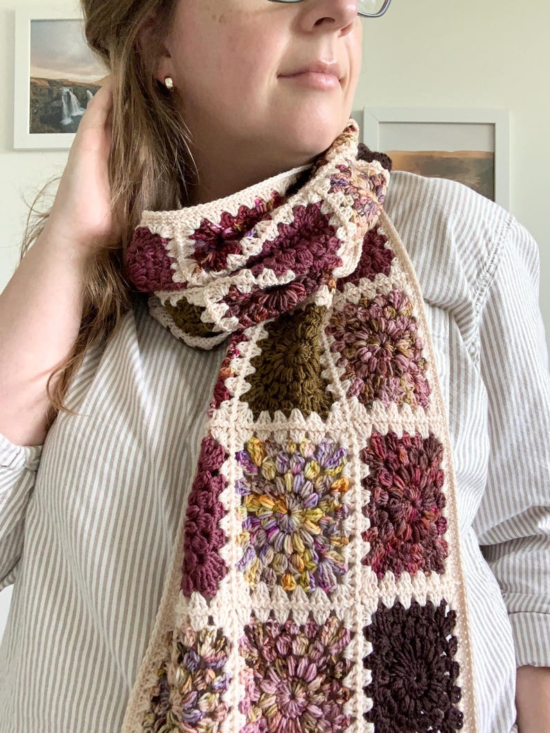 May include: A crocheted scarf with a multi-colored granny square pattern in shades of brown, pink, and green. The scarf is made with a cream-colored yarn.