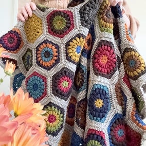 May include: A handmade crochet blanket featuring a repeating pattern of multicolored hexagonal motifs. Each hexagon contains a circular floral design in shades of mustard yellow, deep red, forest green, navy blue, and rust orange, all joined by light beige yarn.