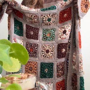 May include: A handmade crochet blanket featuring a patchwork design with square floral motifs. The blanket showcases a palette of earthy tones, including brown, green, orange, and beige, with a gray border. The blanket is held up, displaying its intricate details.