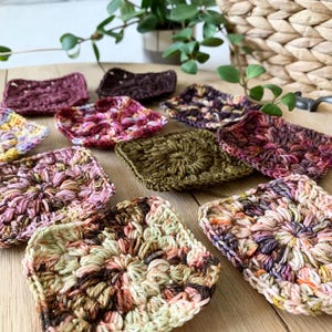 May include: A collection of 10 colorful crocheted squares in various shades of pink, brown, green, and purple. The squares are arranged on a wooden surface.