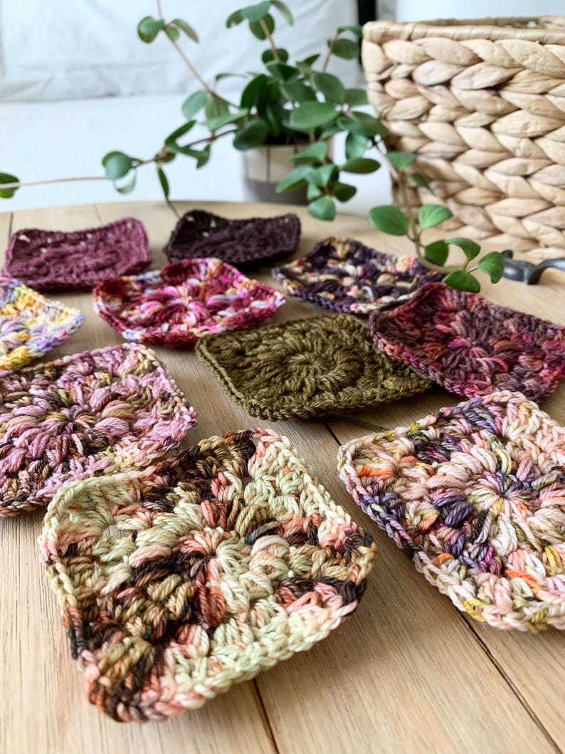 May include: A collection of 10 colorful crocheted squares in various shades of pink, brown, green, and purple. The squares are arranged on a wooden surface.