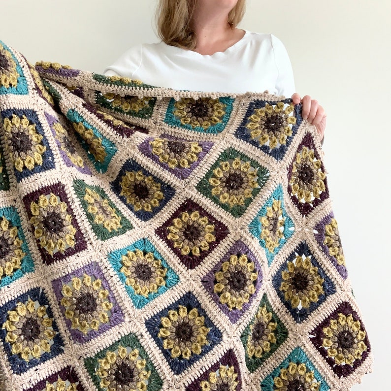May include: A crocheted throw blanket with a sunflower pattern in shades of brown, beige, green, purple, and blue.