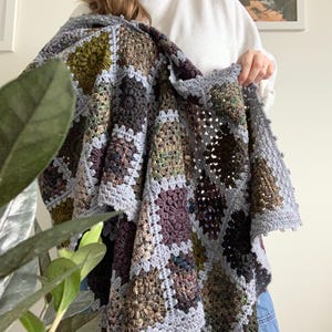 May include: A handmade crochet blanket with a patchwork design. The blanket features squares in shades of grey, green, brown, and purple. The edges are finished with a grey border. The blanket is held up, showcasing its texture and colour variations.