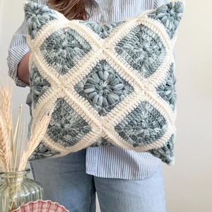 Może przedstawiać: Kwadratowa poduszka szydełkowa z geometrycznym wzorem. Poduszka ma wzór rombu z naprzemiennymi sekcjami jasno-szaro-niebieskiej i kremowej przędzy. Poduszka jest trzymana przed osobą ubraną w niebiesko-białą koszulę w paski i niebieskie dżinsy.