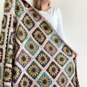 May include: A crocheted throw blanket featuring a sunflower design in shades of brown, green, blue, and purple. The blanket is crafted from textured yarn and has a fringed edge.