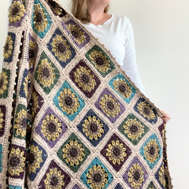 May include: A crocheted throw blanket featuring a sunflower design in shades of brown, green, blue, and purple. The blanket is crafted from textured yarn and has a fringed edge.