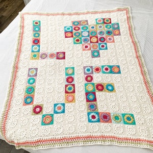 May include: A white crocheted blanket with a colorful flower design spelling out the word "LIFE". The blanket has a crocheted border with pink, green, and white accents.