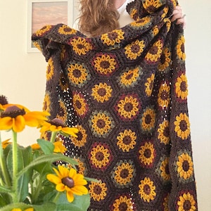 May include: A crocheted blanket with a sunflower pattern. The blanket is predominantly brown, with yellow, orange, and green sunflower designs. The blanket is displayed in a room with natural light.