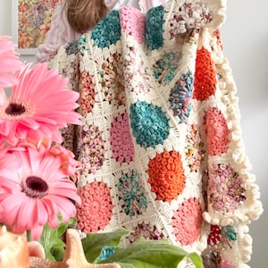 May include: A handmade crochet blanket with a floral design in cream, pink, orange, and teal. The blanket has a scalloped edge. The blanket is a square shape. The blanket is held up in front of a person.