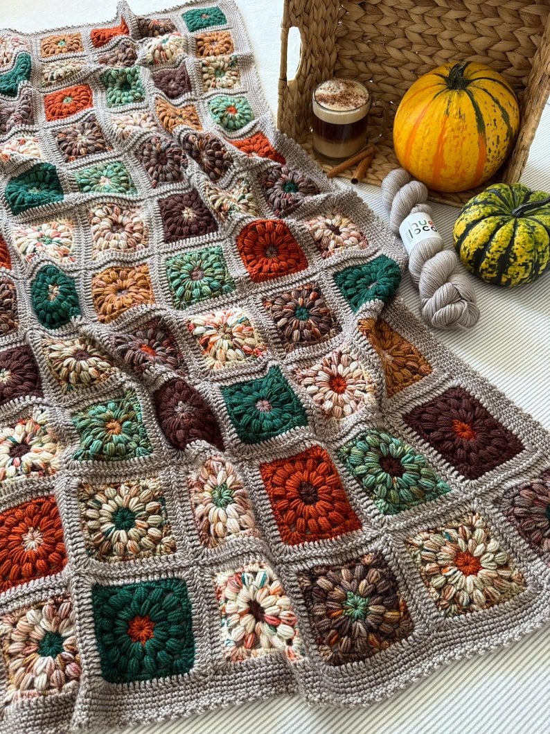 May include: A handmade, square-patterned crochet blanket in a variety of fall colors, including orange, green, brown, and cream. The blanket is draped over a surface, with a basket, pumpkins, and yarn in the background.