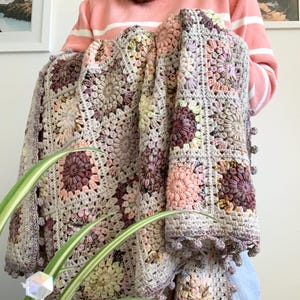 Może przedstawiać: Ręcznie robiony koc szydełkowy w odcieniach szarości, różu i fioletu. Koc ma kwiatowy wzór z teksturowanym wzorem i wykończeniem z pomponów. Koc jest trzymany przez osobę ubraną w różowy sweter.