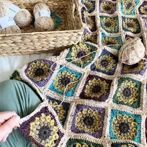 May include: A crocheted blanket with a sunflower pattern in shades of brown, green, yellow, and purple. The blanket is being worked on, with a single square being attached to the rest of the blanket. Balls of yarn are in a wicker basket.