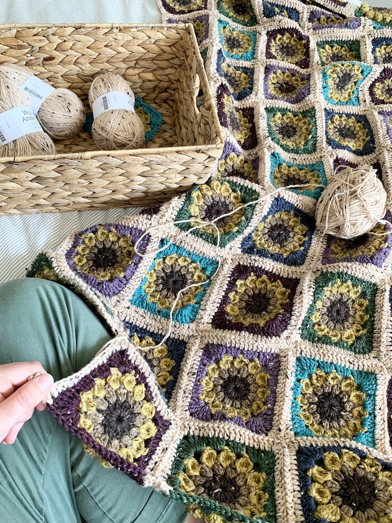 May include: A crocheted blanket with a sunflower pattern in shades of brown, green, yellow, and purple. The blanket is being worked on, with a single square being attached to the rest of the blanket. Balls of yarn are in a wicker basket.