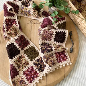 May include: A crocheted scarf with a square pattern in shades of brown, green, and pink. The scarf has a white crocheted border and a fringe.
