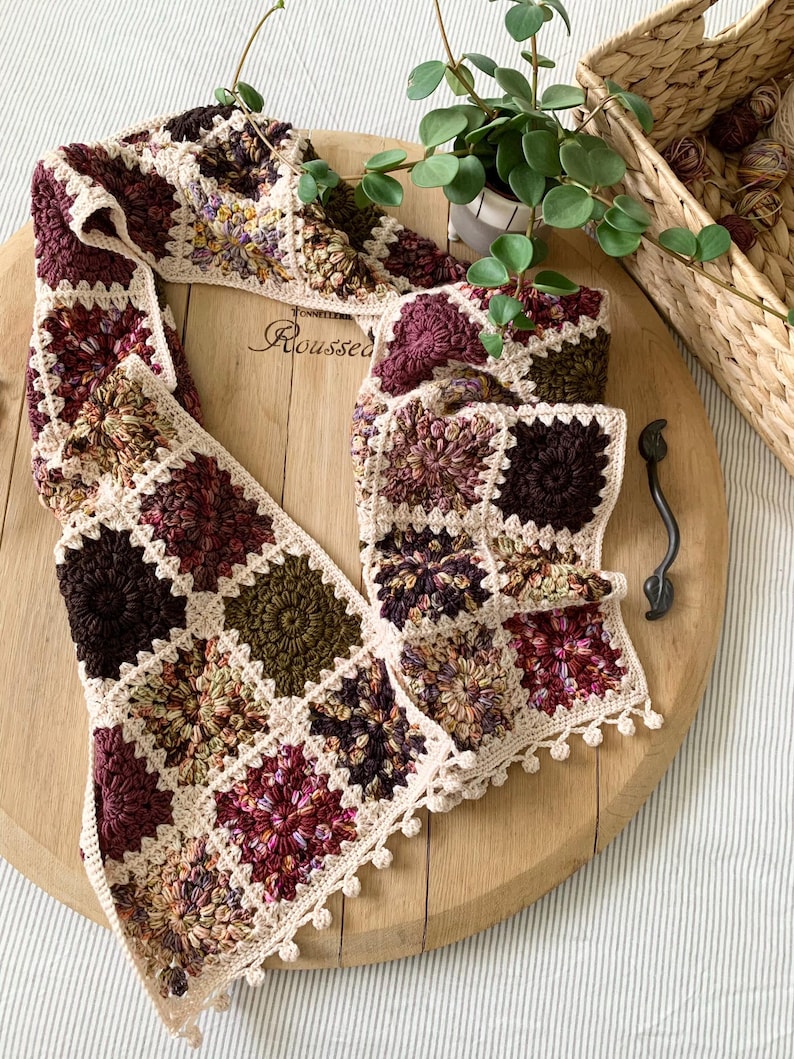 May include: A crocheted scarf with a square pattern in shades of brown, green, and pink. The scarf has a white crocheted border and a fringe.