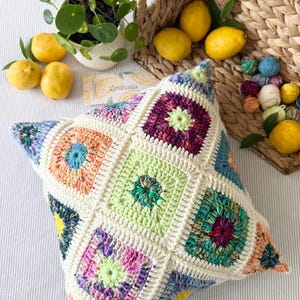 Puede incluir: Un cojín de ganchillo cuadrado que presenta un mosaico de cuadrados de la abuela multicolores unidos por hilo de color crema. Los cuadrados muestran varios tonos vibrantes, incluidos verde lima, morado, naranja y azul, creando un patrón geométrico texturizado.