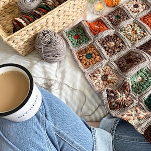 May include: A cozy scene featuring a handmade crochet blanket with floral squares in shades of brown, orange, and green. A basket holds yarn and crochet supplies. A mug of coffee and a small pumpkin complete the autumn-themed setting.