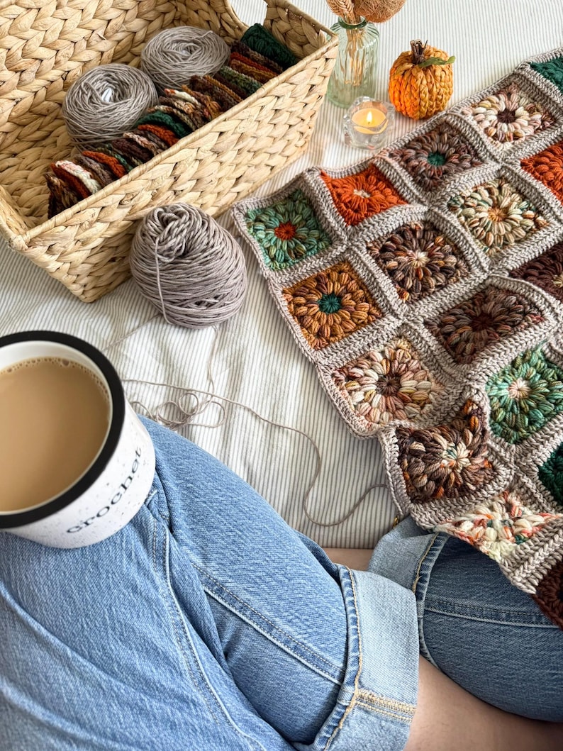 May include: A cozy scene featuring a handmade crochet blanket with floral squares in shades of brown, orange, and green. A basket holds yarn and crochet supplies. A mug of coffee and a small pumpkin complete the autumn-themed setting.