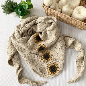 Puede incluir: Una bufanda de ganchillo beige con un patrón moteado y tres diseños de girasol de ganchillo. La bufanda está doblada y colocada sobre una superficie rayada.