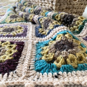 May include: A crocheted blanket featuring a mix of beige, brown, teal and purple colours. The blanket is made up of squares with different patterns. The blanket is lying on a white and grey striped surface.