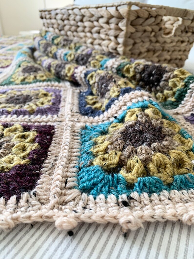May include: A crocheted blanket featuring a mix of beige, brown, teal and purple colours. The blanket is made up of squares with different patterns. The blanket is lying on a white and grey striped surface.