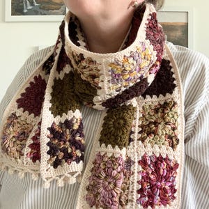 May include: A crocheted scarf with a patchwork design featuring various shades of brown, pink, and green. The scarf has a fringe border and is made with a variety of stitches.