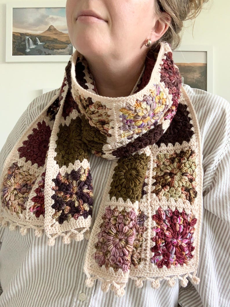 May include: A crocheted scarf with a patchwork design featuring various shades of brown, pink, and green. The scarf has a fringe border and is made with a variety of stitches.