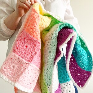 May include: A handmade, colorful crochet blanket featuring a patchwork design. The blanket showcases a vibrant array of pastel colors, including pink, orange, yellow, green, teal, and purple. Each square has a floral pattern, and the blanket is trimmed with a white border.