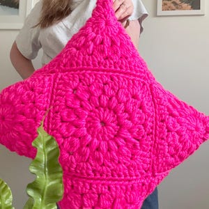 May include: A square-shaped, bright hot pink crochet pillow cover featuring a prominent central sunburst pattern made of thick yarn. The design consists of repeating puff stitches radiating from the center, creating a textured, geometric surface.