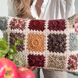 Puede incluir: Un cojín rectangular con un patrón de cuadrados de ganchillo en varios tonos tierra, incluyendo rojo intenso, amarillo mostaza, marrón oscuro y verde salvia, unidos por un borde de hilo color crema en una disposición de cuadrícula.