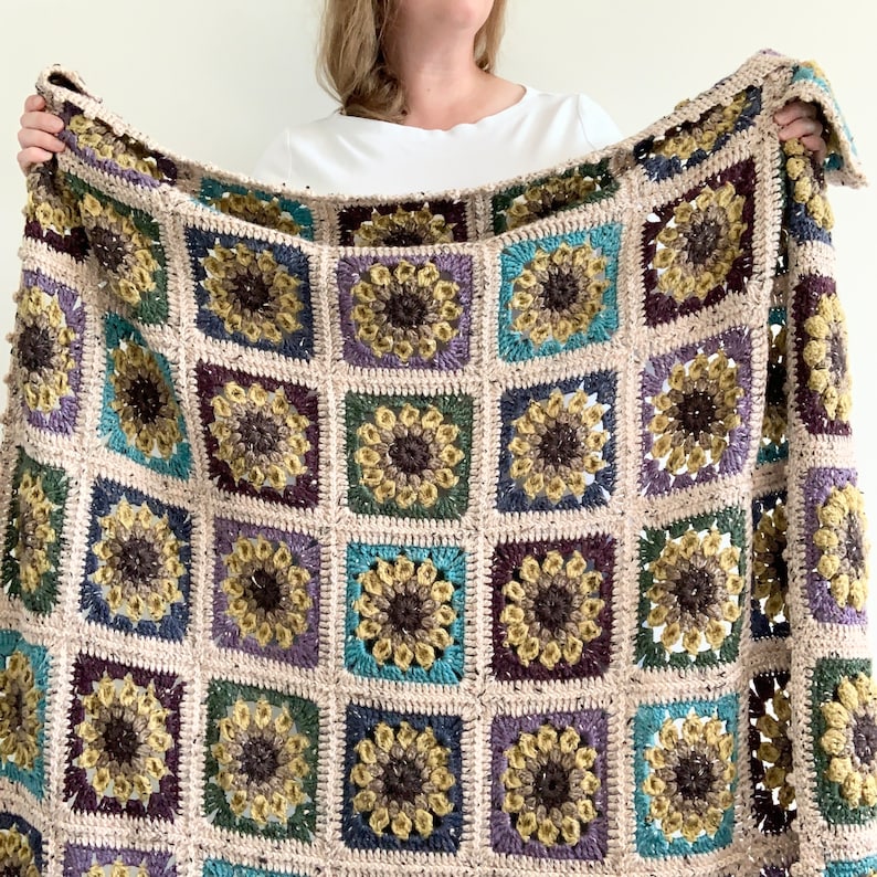 May include: A crocheted throw blanket with a sunflower pattern in shades of brown, beige, purple, teal, and green.