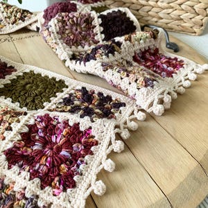 May include: A crocheted scarf with a granny square pattern in shades of brown, green, and pink. The scarf has a white crocheted border with a fringe of white pom-poms.