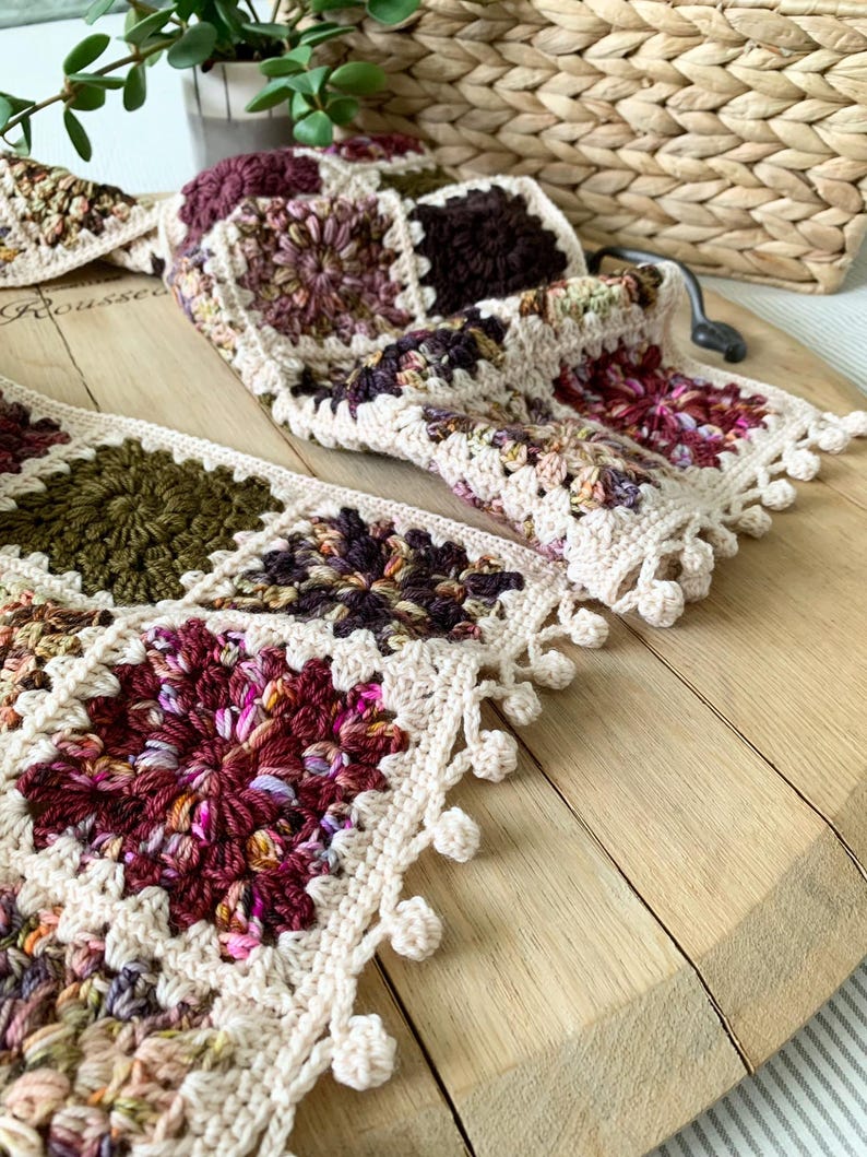 May include: A crocheted scarf with a granny square pattern in shades of brown, green, and pink. The scarf has a white crocheted border with a fringe of white pom-poms.