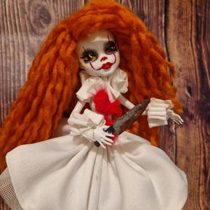 May include: A handmade doll with red and white clown makeup, orange yarn hair, and a white dress. The doll is holding a rusty knife.