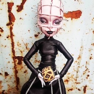 May include: A black leather doll with a grid pattern on its face and a yellow and brown box in its hands. The doll has a black leather outfit and a silver chain around its waist.
