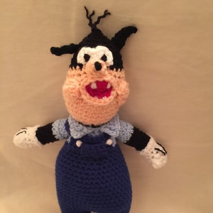 May include: A crocheted plush toy of a cartoon dog wearing blue overalls. The dog has black fur, a white nose, and a big smile.