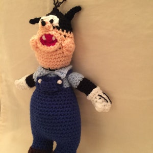 May include: Crocheted plush toy of a cartoon dog wearing blue overalls and a light blue shirt. The dog has black ears, a black nose, and white eyes.