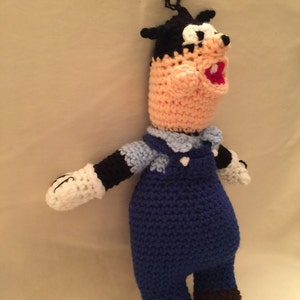 May include: Crocheted plush toy of a cartoon character with black hair, a pink face, and a blue jumpsuit. The toy has white gloves and brown shoes.