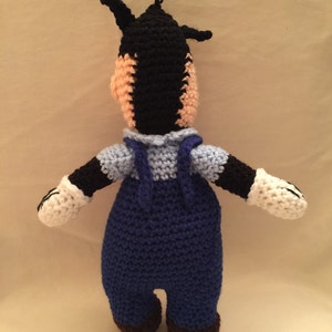 May include: A crocheted stuffed toy of a cartoon character wearing blue overalls. The toy has black fur, a white face, and white gloves.