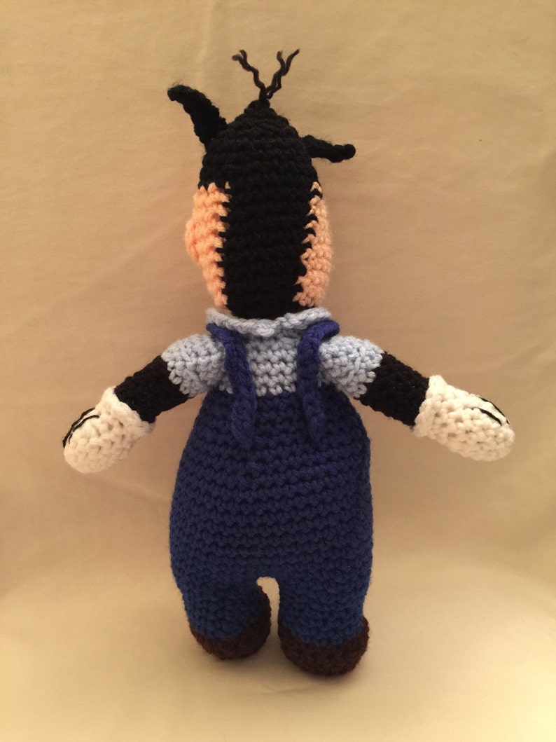 May include: A crocheted stuffed toy of a cartoon character wearing blue overalls. The toy has black fur, a white face, and white gloves.