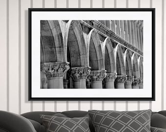 May include: A black and white framed print of an arched architectural structure hangs above a dark gray sofa with patterned pillows. A silver lamp and decorative vases sit on a white side table. The background features a striped wallpaper.