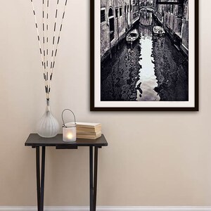 May include: Black and white framed photograph of a Venetian canal scene. A small black table holds a vase with decorative sticks, a candle lantern, and a stack of books. The photograph depicts a canal with buildings on either side.