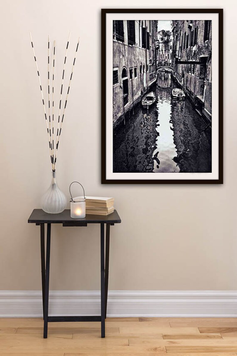 May include: Black and white framed photograph of a Venetian canal scene. A small black table holds a vase with decorative sticks, a candle lantern, and a stack of books. The photograph depicts a canal with buildings on either side.