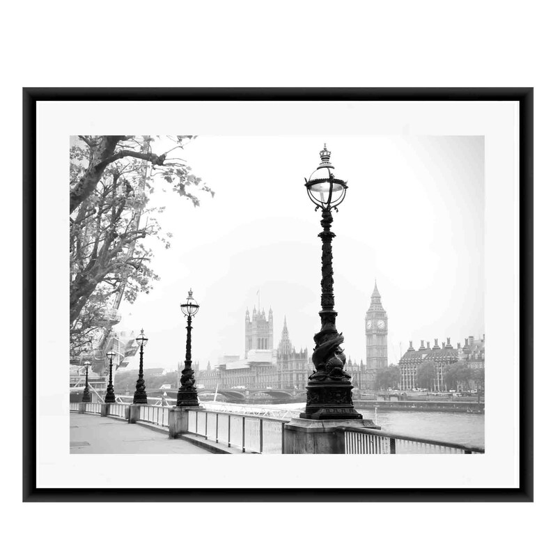 May include: A black and white photograph of a street scene in London, England. The photo shows a row of lampposts along a sidewalk, with the Houses of Parliament and Big Ben in the background. The river Thames is visible in the distance.