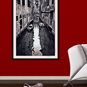 May include: Black and white framed print of a Venetian canal scene. The image shows a narrow waterway with buildings on either side, a bridge, and boats. The print is hanging on a red wall, with a pair of white high-heeled sandals on the floor.