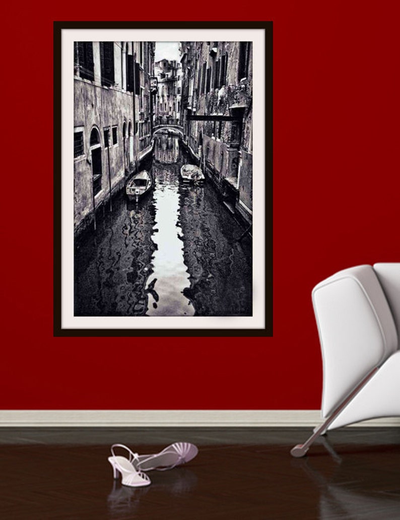 May include: Black and white framed print of a Venetian canal scene. The image shows a narrow waterway with buildings on either side, a bridge, and boats. The print is hanging on a red wall, with a pair of white high-heeled sandals on the floor.
