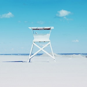 May include: A white lifeguard stand on a sandy beach with a blue sky and white clouds in the background.