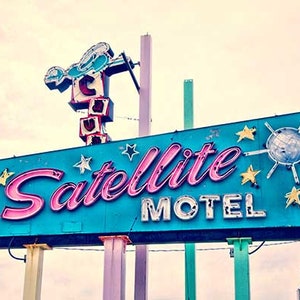 May include: A vintage neon sign for a motel called "Satellite Motel". The sign is blue with pink neon lettering and features a stylized starburst with a disco ball in the center.