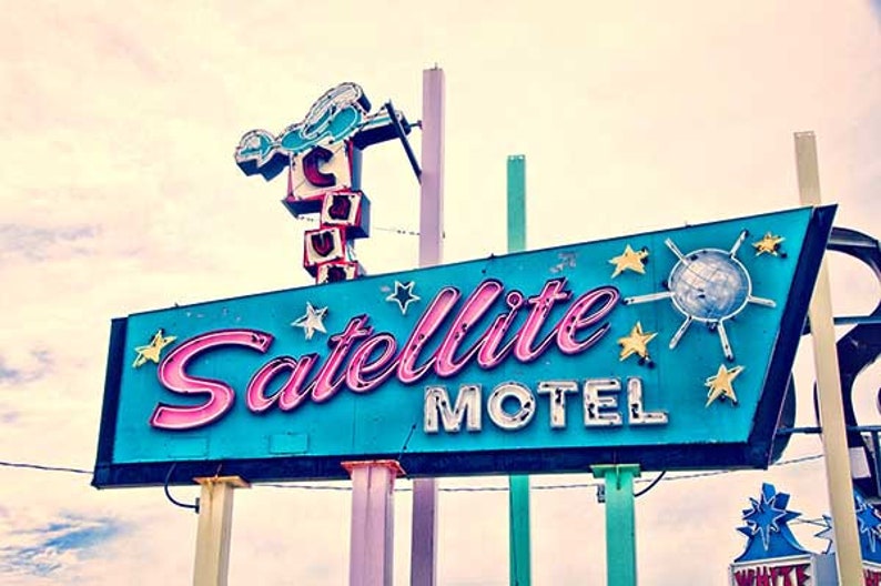 May include: A vintage neon sign for a motel called "Satellite Motel". The sign is blue with pink neon lettering and features a stylized starburst with a disco ball in the center.